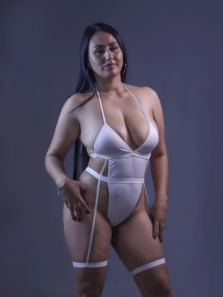 ValentinaJaramilloo with long black hair, wearing a black top and posing with her hands up near her face in a room with wooden shelving.