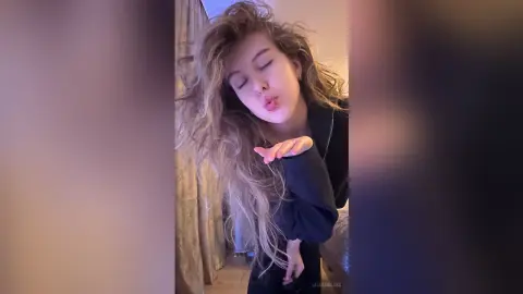 Blonde woman in a black sweater blowing a kiss with messy hair in a cozy indoor setting.
