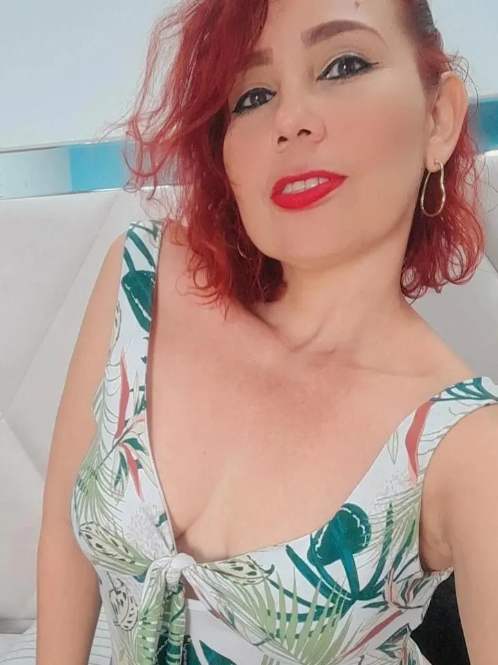nayasex199 smiles in a white floral print tank top with her red hair and gold hoop earrings.