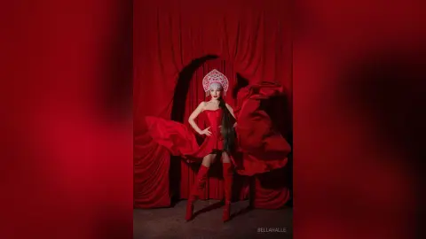 Elegant woman in a red flowing gown and ornate silver headpiece posing against red velvet curtains.