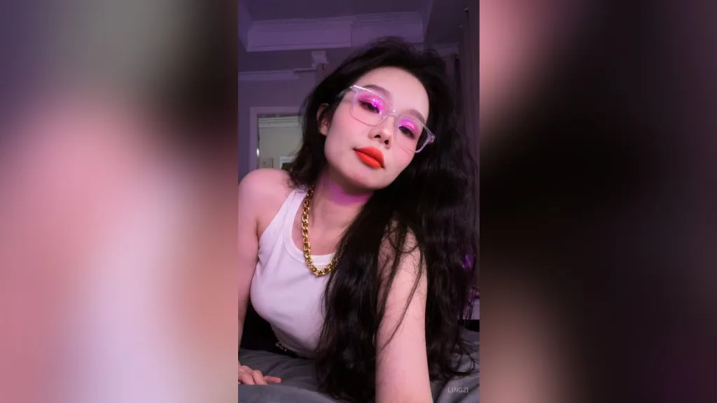 lingzi — lingzi wears a white tank top and gold chain necklace while posing in a room with pink lighting.