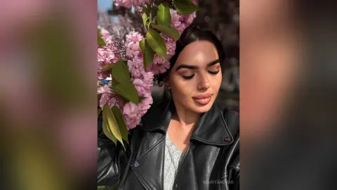 Beautiful woman in a black leather jacket posing near blooming pink flowers in soft sunlight.