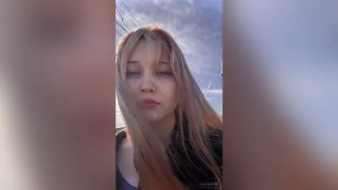 EloiseGow looks into the camera with long blonde hair against a cloudy sky background.
