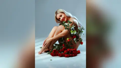 Anita poses naked on a white floor, surrounded by red roses and green leaves that partially cover her body.