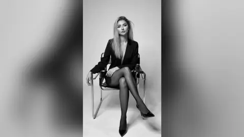 Elegant woman in a black blazer and heels sitting on a chair in a minimalist studio setting.