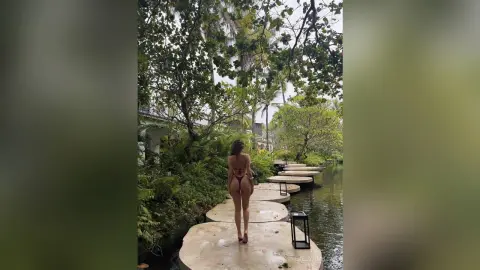 Woman in a brown bikini walking on a stone path through a lush tropical garden by a serene pond.