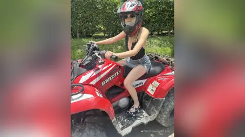 Young woman in black tank top and denim shorts riding a red Yamaha Grizzly ATV in a lush green outdoor setting.