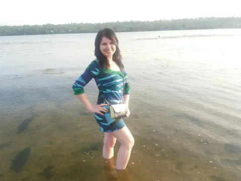 Smiling woman in a blue patterned summer dress standing in shallow lake water on a sunny day.