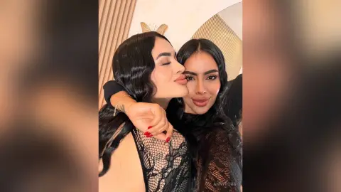 Two smiling women in black lace outfits sharing a joyful embrace in a modern, brightly lit room.