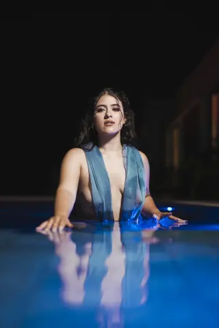 Young woman in a blue wrap relaxing in a glowing swimming pool at night under moody lighting.