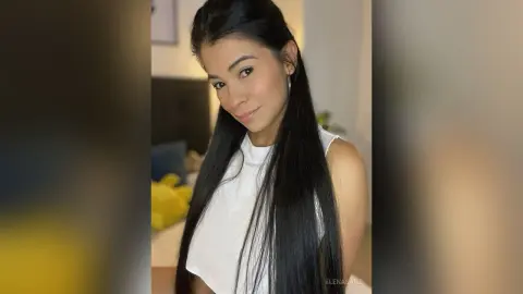 Smiling young woman with long dark hair wearing a white sleeveless top in a bright indoor setting.