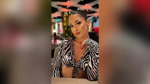 Young woman in a black and white zebra print crop top posing in a dimly lit restaurant setting.
