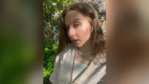 Young woman in a white checkered blazer and pearl necklace posing outdoors in sunny garden setting.