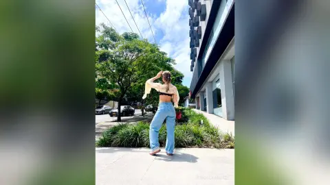 Young woman in light blue jeans and a white crop top standing on a sunny city sidewalk near green trees.