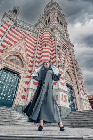 Nun in a long grey habit standing on cathedral steps before a grand red and white striped church under cloudy skies.