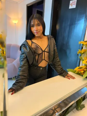 Smiling young woman in a black leather jacket and sheer bodysuit posing in a modern living room.