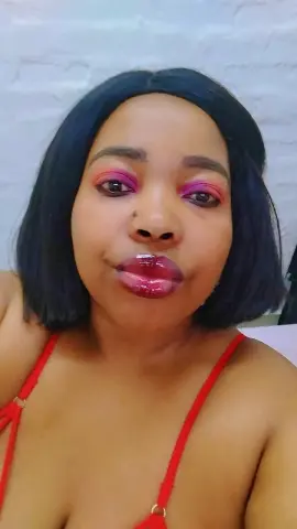 SexyChubbyxx29 wearing red lingerie and pink eyeshadow, posing with a pouty lip in front of a white brick wall.