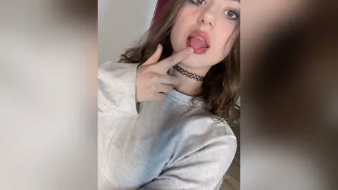 AriaLush pouts with her tongue out and finger touching her lip, wearing a grey long-sleeve shirt and a black choker.