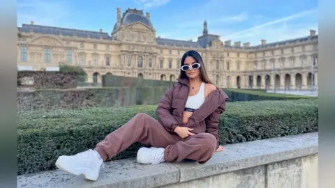 TefyRusso wears a brown puffer jacket and matching sweatpants with a white sports bra, sitting on a stone ledge in front of a large palace building.