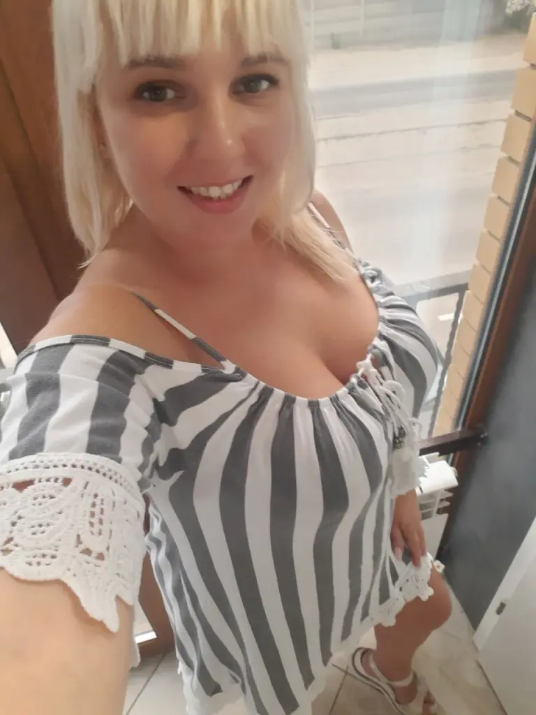 Djessika — Smiling blonde woman in a striped off-the-shoulder summer top relaxing by a bright window.