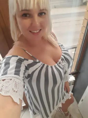 Smiling blonde woman in a striped off-the-shoulder summer top relaxing by a bright window.