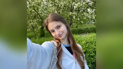 Smiling young woman in a white hoodie taking a selfie outdoors near blooming spring trees.