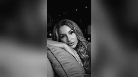 Portrait of a smiling woman with long wavy hair resting on a pillow in a moody black and white setting.