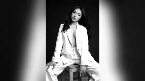 Elegant woman in a white tailored suit sitting on a stool in a moody black and white studio portrait.