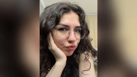Young woman with curly hair and glasses making a playful pouty face in a cozy indoor setting.