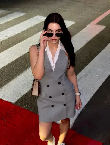 Stylish woman in a grey sleeveless waistcoat dress and sunglasses standing on a city crosswalk at night.