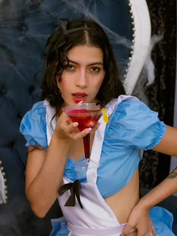 SusanaSanchez20 wears a blue and white Alice in Wonderland outfit while holding a red cocktail in a martini glass.