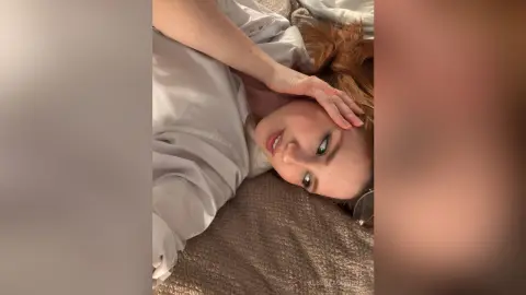 AlbertaCorrell lies on a bed with her red hair spread out, wearing a white shirt and looking into the camera.