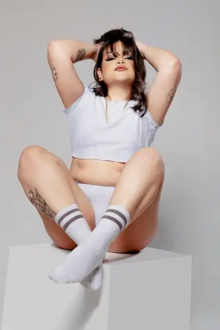 Young woman in white crop top and socks sitting on a white block in a minimalist studio setting.