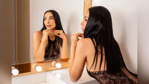 Elegant woman in a black lace bodysuit looking into a mirror in a bright, modern room.