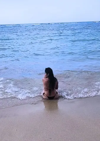 Young woman in a black bikini sitting on a sandy beach watching ocean waves under a blue sky.