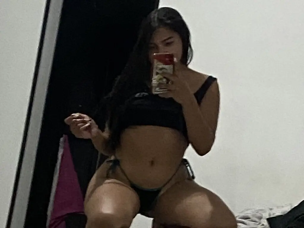 Young woman in black crop top and bikini posing for a mirror selfie in a dimly lit room.