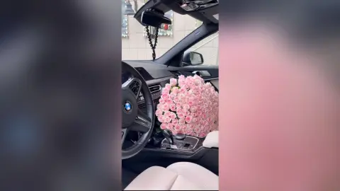 SerenaBliss sits in a car seat next to a large, dense bouquet of pink roses on the passenger side.