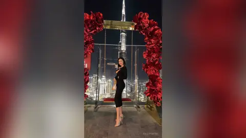 Elegant woman in a black evening dress sitting on a swing with red floral decor overlooking the Dubai skyline.