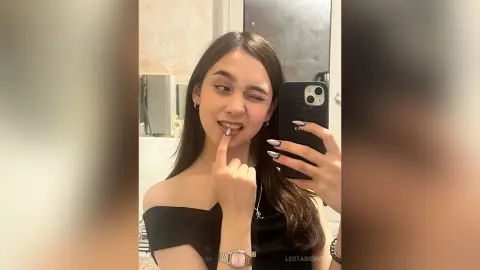 Young woman with long dark hair winking and taking a mirror selfie in a black off-the-shoulder top.