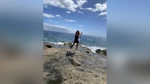 Stylish woman in a black romper and white shirt standing on rocky cliffs by the blue ocean under a sunny sky.