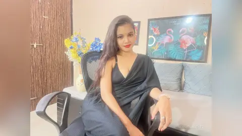 AdrianaReyez poses in a black low-cut dress with long dark hair, sitting in an office chair against a light wall with a framed flamingo print.
