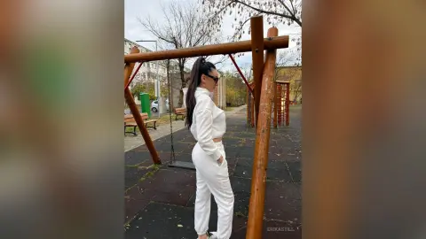 Stylish woman in white loungewear standing by a wooden swing set in a quiet urban park on an overcast day.