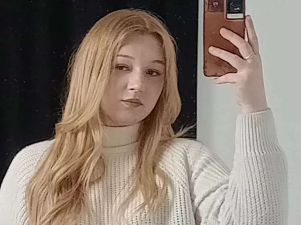 Blonde woman in a cozy white ribbed turtleneck sweater taking a casual mirror selfie at home.