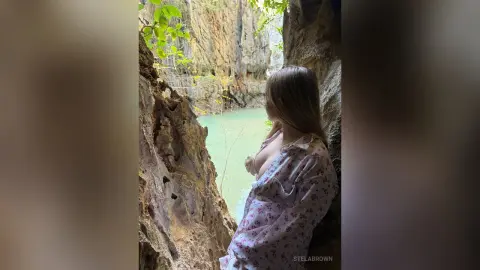 Blonde woman in a white floral dress looking out from a rocky canyon over a turquoise river in a serene forest.