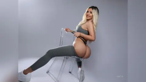 Anita sits sideways on a clear acrylic chair wearing a grey sports bra and matching leggings, showing off her bare buttocks and blonde hair.