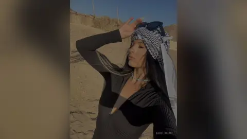 Stylish woman in a black long-sleeve top and patterned headscarf posing on a sunny desert beach.