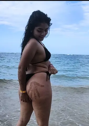 Smiling young woman in a black bikini standing on a sunny beach by the ocean waves.