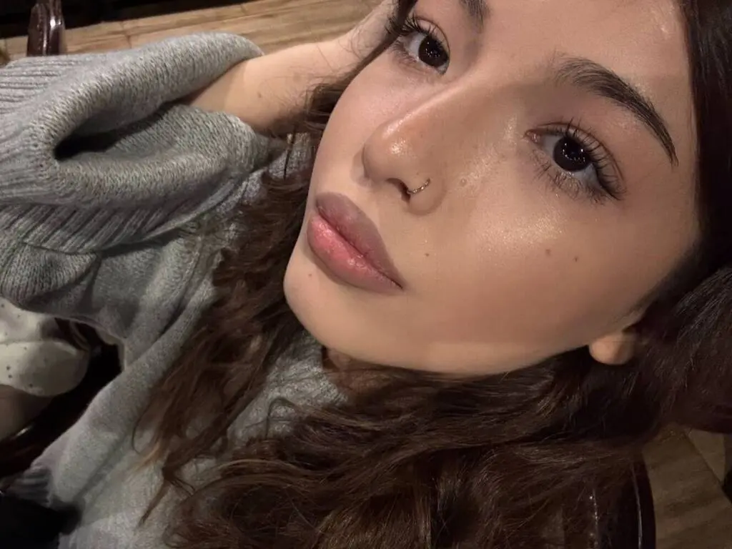 Close-up portrait of a young woman with wavy brown hair wearing a cozy grey sweater in soft indoor lighting.