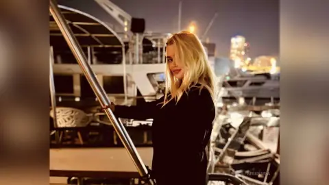 MilenaBia stands on a boat deck at night, wearing a black long-sleeved top with her blonde hair down.