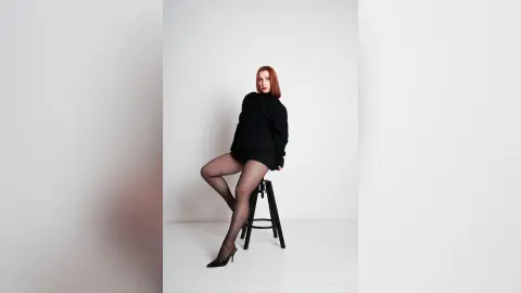 AryaSin poses sitting on a black stool wearing a black long-sleeve mini dress and sheer black tights with black high heels against a white background.